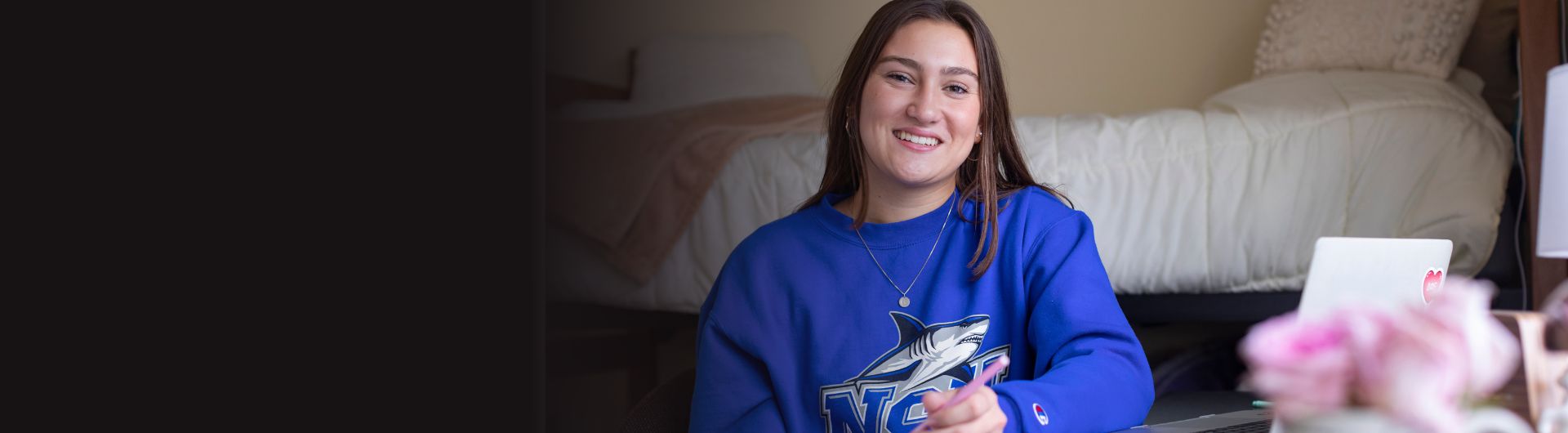 A student wearing an NSU shark shirt is writing on a notepad in her room with a bed in the background.