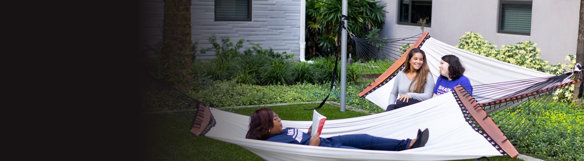 Two hammocks occupied by three students. One student is comfortably stretched out while reading a book. The other two are sitting, chatting, and laughing.
