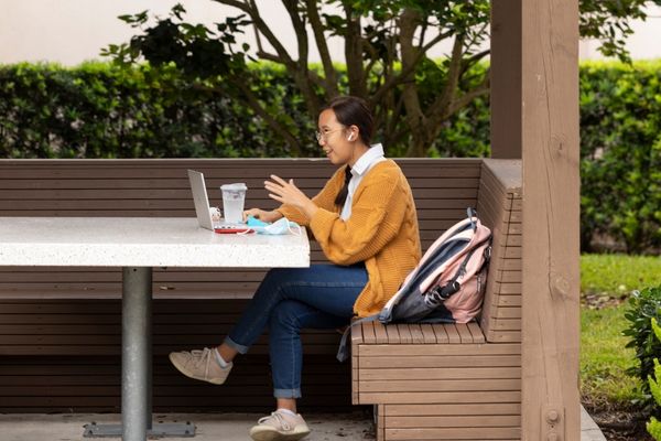 An NSU student is sitting in an outdoors seating area and is engaged in conversation with someone via her laptop and airpods.