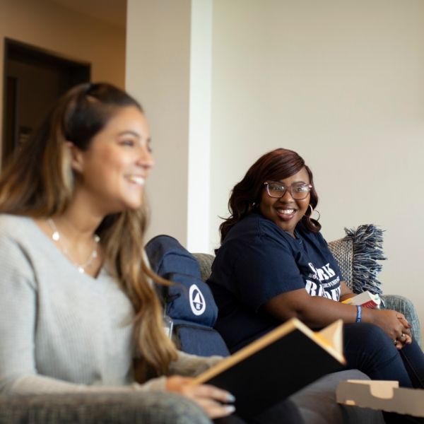 Two smiling and laughing NSU students seated on a gray couch. The one in the front is slightly blurry, but you can see that she’s holding an open book. The in-focus student further from the camera is looking directly into the camera.